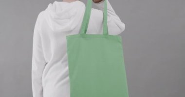 Beyaz kapüşonlu beyaz kadın gri arka planda yeşil çanta tutuyor. Kopya alanı, ağır çekim. Moda, tasarım, kıyafetler ve kumaş.