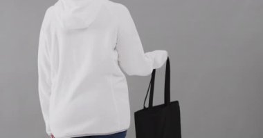 Beyaz kapüşonlu beyaz bir kadın gri arka planda siyah bir çanta tutuyor. Kopya alanı, ağır çekim. Moda, tasarım, kıyafetler ve kumaş.