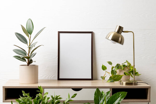 Black empty frame with copy space, lamp and plants in pots on table against white wall. Mock up frame template, interior design and decoration.