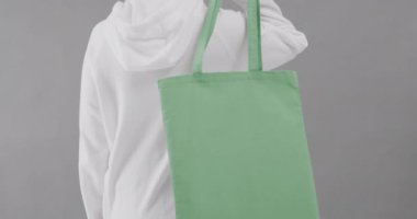 Beyaz kapüşonlu beyaz kadın gri arka planda yeşil çanta tutuyor. Kopya alanı, ağır çekim. Moda, tasarım, kıyafetler ve kumaş.