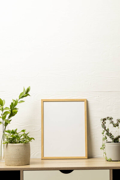 Vertical of empty wooden frame with copy space and plants in pots on desk against white wall. Mock up frame template, interior design and decoration.