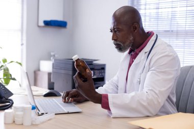 Afrika kökenli Amerikalı erkek doktor elinde ilaçla masada oturuyor ve doktorun ofisinde dizüstü bilgisayar kullanıyor. Hastane, ilaç, sağlık ve iletişim.