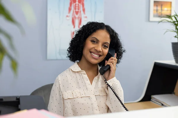 stock image Portrait of happy biracial medical receptionist sitting at reception desk and talking on telephone. Hospital, medicine and healthcare.