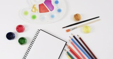 Ağır çekimde, beyaz arka planda defter, boya, fırça ve boya kalemlerinin genel görünümü. Kırtasiye, yaratıcılık, öğrenme, okul ve eğitim kavramı.