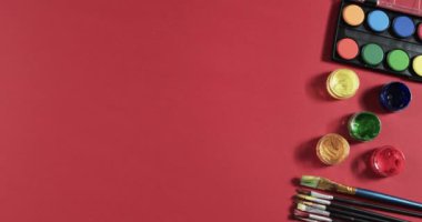 Boyaların ve fırçaların yukarıdan görünüşü kırmızı arkaplanda, fotokopi alanı ile, yavaş çekimde. Kırtasiye, yaratıcılık, öğrenme, okul ve eğitim kavramı.