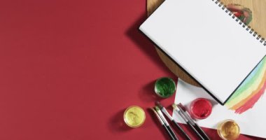 Not defterinin yukarıdan görünüşü, kırmızı arkaplanda fotokopi alanı ile yavaş çekimde boya ve fırçalar. Kırtasiye, yaratıcılık, öğrenme, okul ve eğitim kavramı.