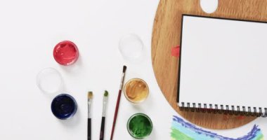 Not defterinin yukarıdan görünüşü, beyaz arka planda fırçalar ve boyalar fotokopi alanı ile, yavaş çekimde. Kırtasiye, yaratıcılık, öğrenme, okul ve eğitim kavramı.