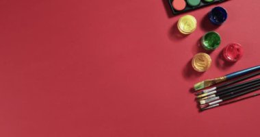 Boyaların ve fırçaların yukarıdan görünüşü kırmızı arkaplanda, fotokopi alanı ile, yavaş çekimde. Kırtasiye, yaratıcılık, öğrenme, okul ve eğitim kavramı.