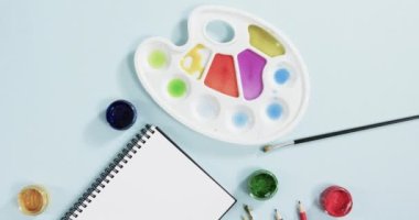 Defterin yukarıdan görünüşü, fırçalar ve boyalar mavi arka planda, yavaş çekimde. Kırtasiye, yaratıcılık, öğrenme, okul ve eğitim kavramı.