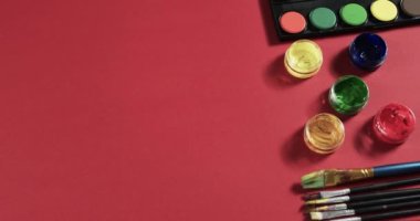 Boyaların ve fırçaların yukarıdan görünüşü kırmızı arkaplanda, fotokopi alanı ile, yavaş çekimde. Kırtasiye, yaratıcılık, öğrenme, okul ve eğitim kavramı.
