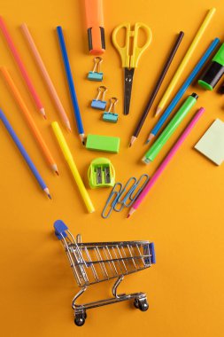 Minyatür alışveriş vagonunu turuncu arka planda okul malzemeleriyle kapatın. Okula dönüş, alışveriş, okul malzemeleri, öğrenme, okul ve eğitim kavramı.