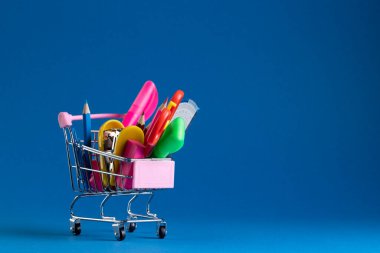 Minyatür alışveriş arabasını okul malzemeleriyle kapatın ve mavi arka planda fotokopi alanı oluşturun. Okula dönüş, alışveriş, okul malzemeleri, öğrenme, okul ve eğitim kavramı.