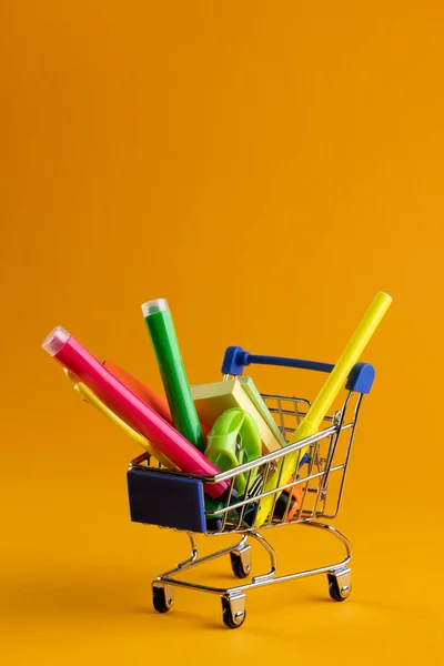 stock image Close up of miniature shopping trolley with school materials and copy space on orange background. Back to school, shopping, school materials, learning, school and education concept.