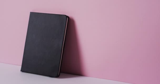 遅い動きでピンクの背景にコピースペースと壁にもたれて閉じられた黒い本のクローズアップ — ストック動画