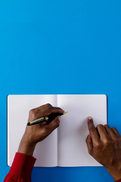 Hands of biracial man holding pen and writing in notebook with copy space on blue background. Literature, writing, leisure time and books.