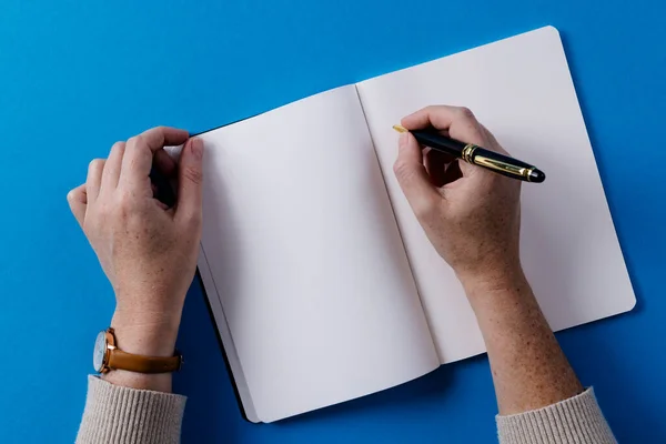 Hands of caucasian woman holding pen and writing in notebook with copy space on blue background. Literature, writing, leisure time and books.