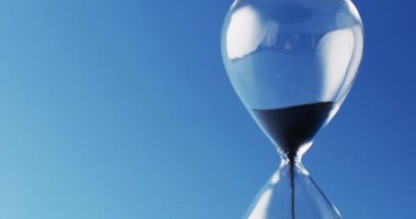 Video of close up of hourglass with black sand pouring with copy space on blue background. Time and time keeping concept.