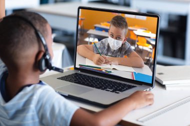 Arkadan Afro-Amerikan bir çocuğun evde beyaz bir çocukla görüntülü konuşma yaparken dizüstü bilgisayar kullanması. Uzak öğrenim çevrimiçi eğitim kavramı