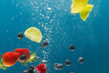 Böğürtlen, limon ve limon dilimlerinin mavi arka planda fotokopi alanı olan suya düşüşünü kapat. Meyve, vejetaryen yemeği ve renk konsepti.