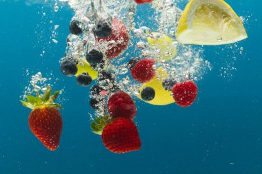 Böğürtlen, limon ve limon dilimlerinin mavi arka planda fotokopi alanı olan suya düşüşünü kapat. Meyve, vejetaryen yemeği ve renk konsepti.