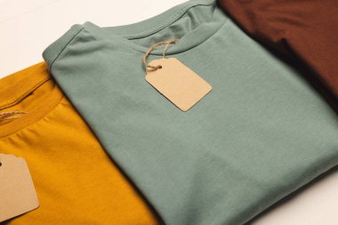 Beyaz tahta arka planda etiketli ve kopyalama alanı olan üç tişörtten oluşan düz yatağı kapatın. Moda, kıyafetler, renk ve kumaş konsepti.