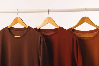 Elbise korkuluklarından sarkan askılarda üç kahverengi tişört ve beyaz arka planda fotokopi alanı. Moda, kıyafetler, renk ve kumaş konsepti.