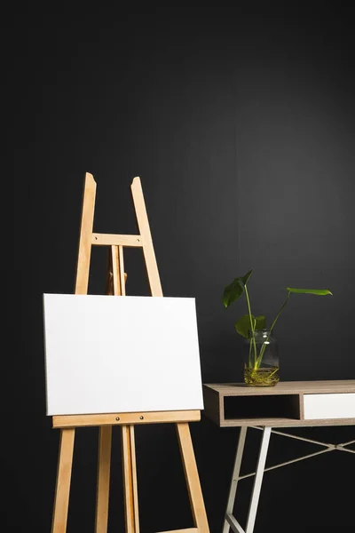 木製のイーゼルに白いキャンバスがあり 黒い背景に机と植物が付いているスペースをコピーします サイネージ ライティングスペース 広告コンセプト — ストック写真