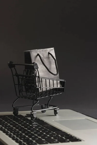 Vertical image of laptop and trolley with bag and copy space on black background. Cyber monday, cyber shopping, retail, technology, electronic device and communication concept.