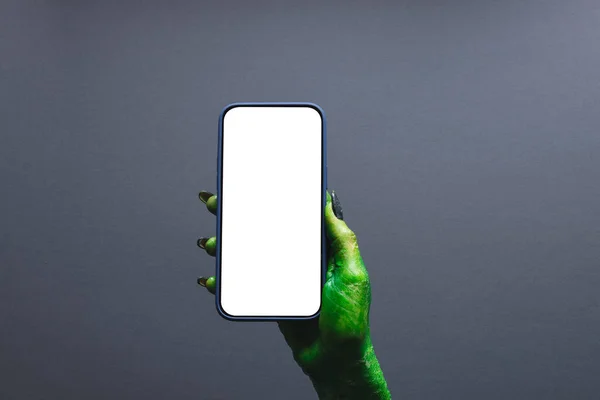 Green monster hand holding smartphone with copy space on grey background. Halloween, tradition and celebration concept.