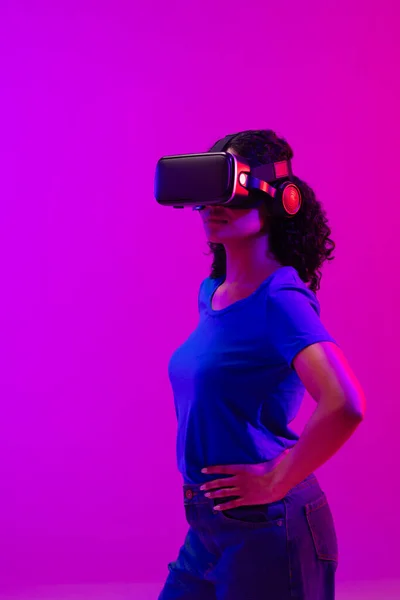 ネオンピンクのヘッドセットを紫色の背景にした女性の垂直画像 テクノロジー デジタルネットワーク グローバルな接続 通信メタバース — ストック写真