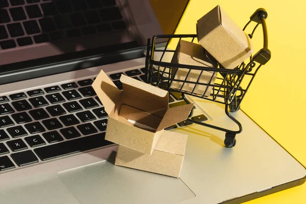 Laptop with copy space and trolley with boxes on yellow background. Cyber shopping, retail, technology, electronic device and communication concept.
