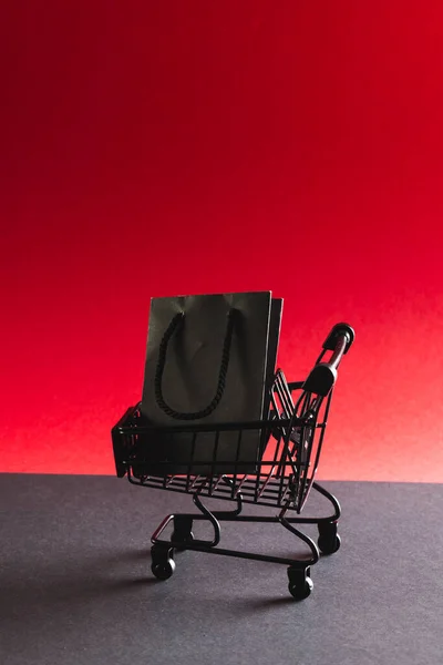 Black gift bag in shopping trolley with copy space on red background. Black friday, cyber monday, shopping, cyber shopping, sales, retail and shipping concept.