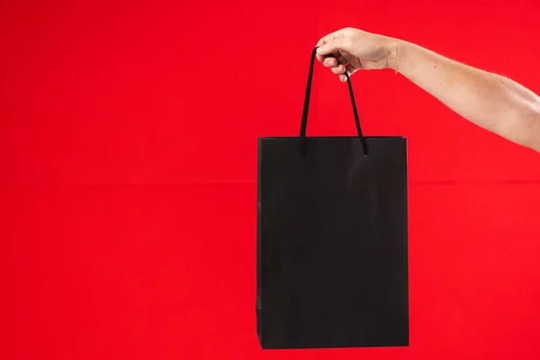 Hand of caucasian woman holding black gift bag with copy space on red background. Black friday, cyber monday, shopping, cyber shopping, sales, retail and shipping concept.