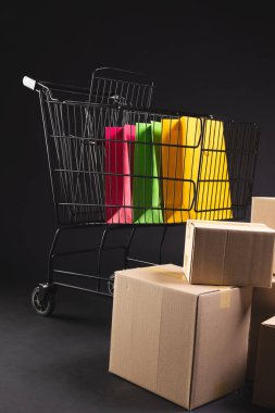 Çantaları, kutuları ve siyah arkaplanı olan alışveriş arabasının dikey görüntüsü. Siber Pazartesi, Kara Cuma, çevrimiçi alışveriş, nakliye ve küresel bağlantılar kavramı.