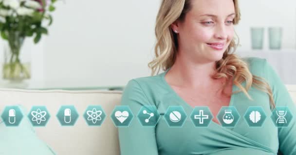 Медичні Значки Над Щасливою Вагітною Білою Жінкою Дивані Допомогою Ноутбука — стокове відео