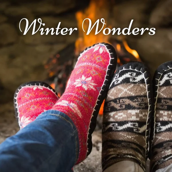 Composite of winter wonders text over man and woman\'s feet in christmas socks by fireplace. Winter, christmas, seasons and celebration concept digitally generated image.