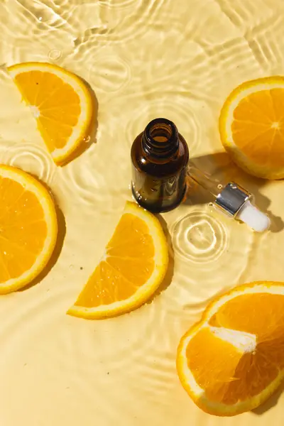Vertical image of bottle with pipette and orange slices in water, copy space on orange background. Health and beauty, make up and beauty concept.