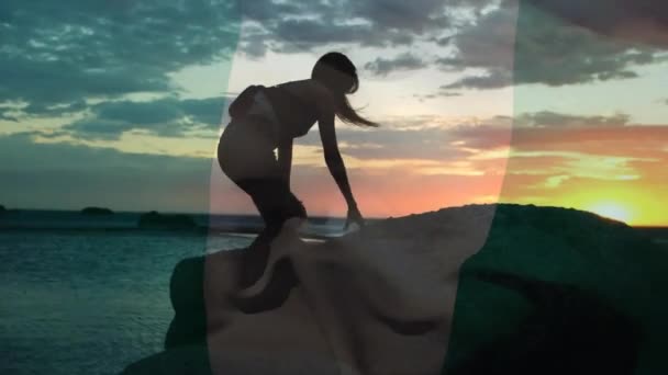 幸せな白人女性の上にナイジェリアの旗のアニメーション 岩の上に登り 日没のビーチを見ます ナイジェリア 愛国心 デジタル生成されたビデオ — ストック動画