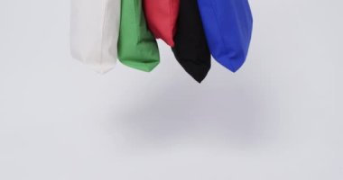 Beyaz, yeşil, kırmızı, siyah ve mavi renkli çantalardan oluşan bir kolleksiyon düz bir arkaplanda asılı duruyor. Bu yeniden kullanılabilir poşetler plastik alışveriş torbalarına çevre dostu bir alternatif teşkil ediyor ve sürdürülebilir uygulamaları teşvik ediyor.