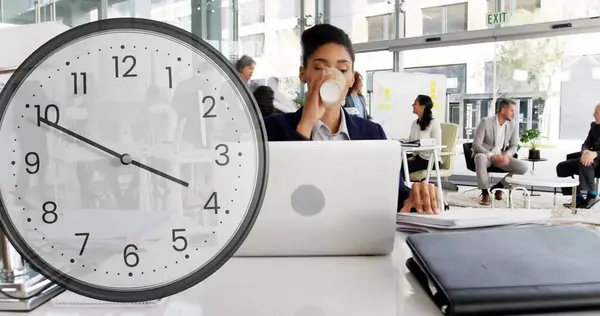 Image of clock ticking over biracial businesswoman in office. Global finance, business, connections, computing and data processing concept digitally generated image.