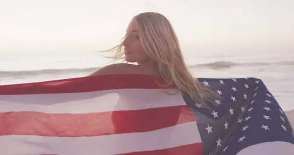 Image of independence day text over smiling woman wrapped in american flag on beach. usa patriotism and democracy concept digitally generated image.