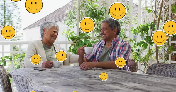 Image of smiling emojis over senior couple drinking coffee at home. global networking, connection and communication concept digitally generated image.
