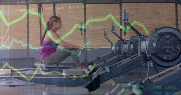 Image of data on graph over biracial woman training on rowing machine at gym. Fitness, exercise, strength, data, digital interface and technology digitally generated image.