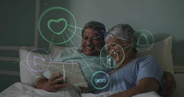 Image of network of connections with icons over senior couple using tablet in bed at home. global social media networking, connection and communication concept digitally generated image.