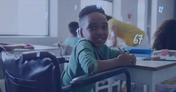 Image of numbers over happy african american schoolboy in wheelchair at desk in class. Disability, inclusivity, school, education, childhood and learning, digitally generated image.