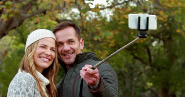 Kafkasyalı genç bir çiftin arka plandaki parkta selfie çubuğuyla selfie çekerken görüntüsü.