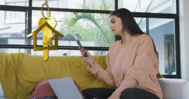 Altın ev anahtarı ve anahtarlığın animasyonu evde akıllı telefon kullanan mutlu çift ırklı kadının üzerinde. Ev, ev, mülk, mülkiyet ve dijital olarak üretilen yaşam tarzı konsepti.