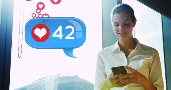 一个白人女人靠近窗户 笑着用手机发短信 在她旁边的前景是一个数字图像的消息泡沫与心脏图标增加计数和图标向上飞 — 图库照片