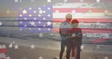 Amerikan bayrağının animasyonu, yıldızlar ve çeşitli çiftlerin güneşli sahilde kucaklaşması. Amerika, yaz, tatiller, romantizm, vatanseverlik ve kutlama, dijital olarak üretilen video.