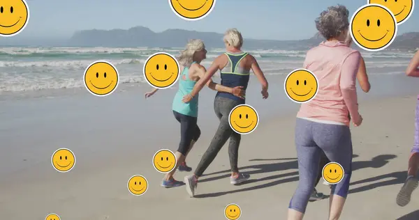 Image of smiley face symbols, over women running on beach. positive feelings and wellbeing social media concept, digitally generated image.